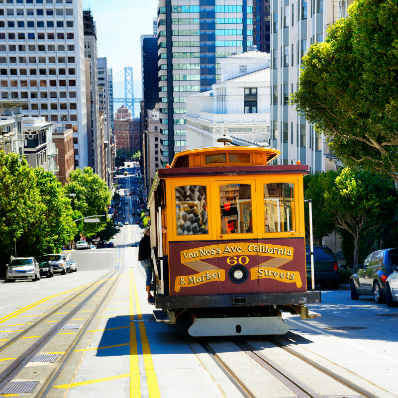 A cable car travels along a hilly San Francisco street, with the Bay Bridge visible in the distance.