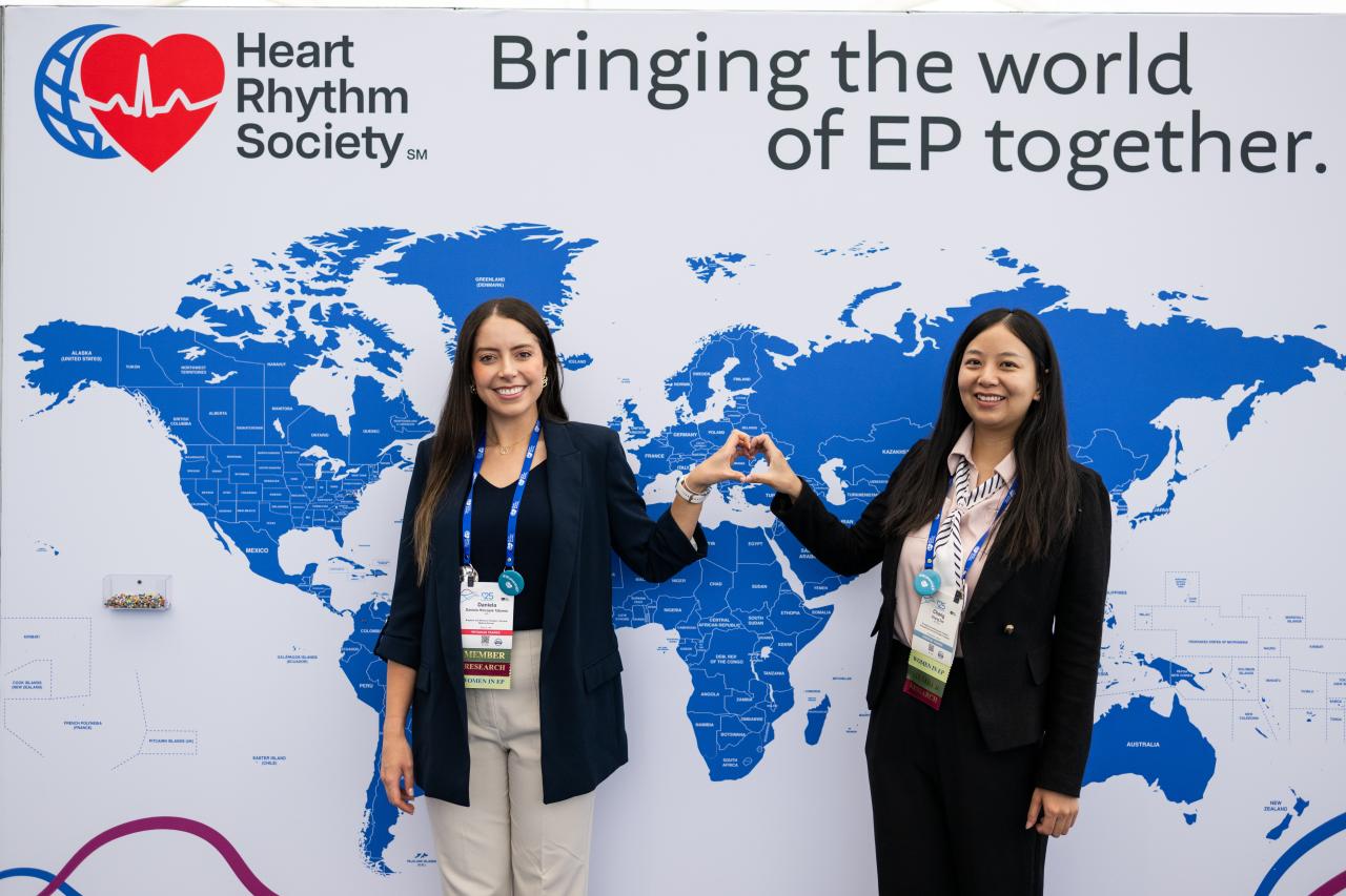 two young heart rhythm attendees making a heart with their hands in front of a world map with the words "bringing the world of EP together"